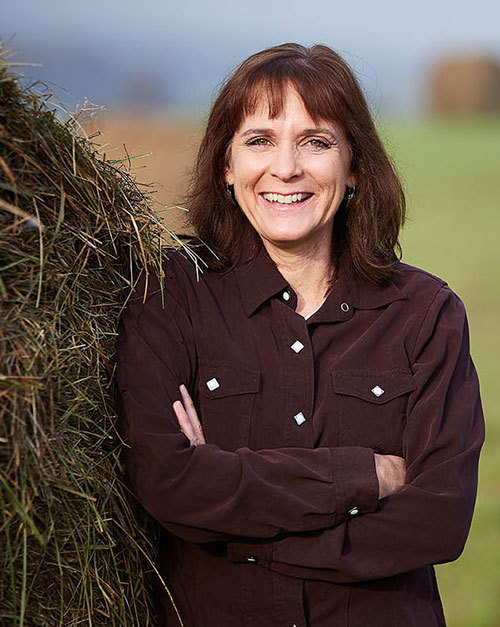 Barb Gleim standing beside a stack of hay bales in front of a farm field.